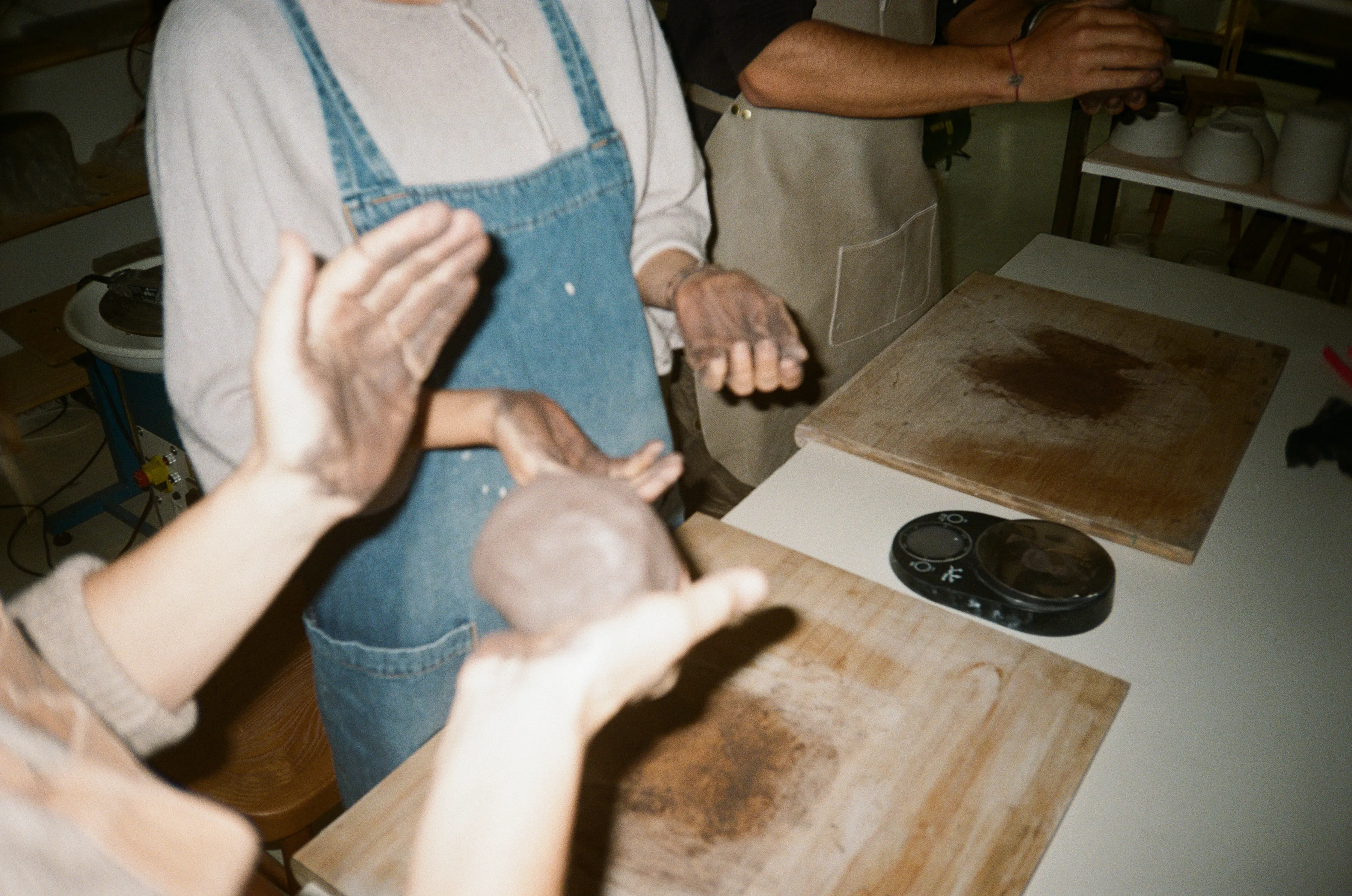 Manos de alumnos amasando bolas de barro en la mesa del taller, con el torno al fondo