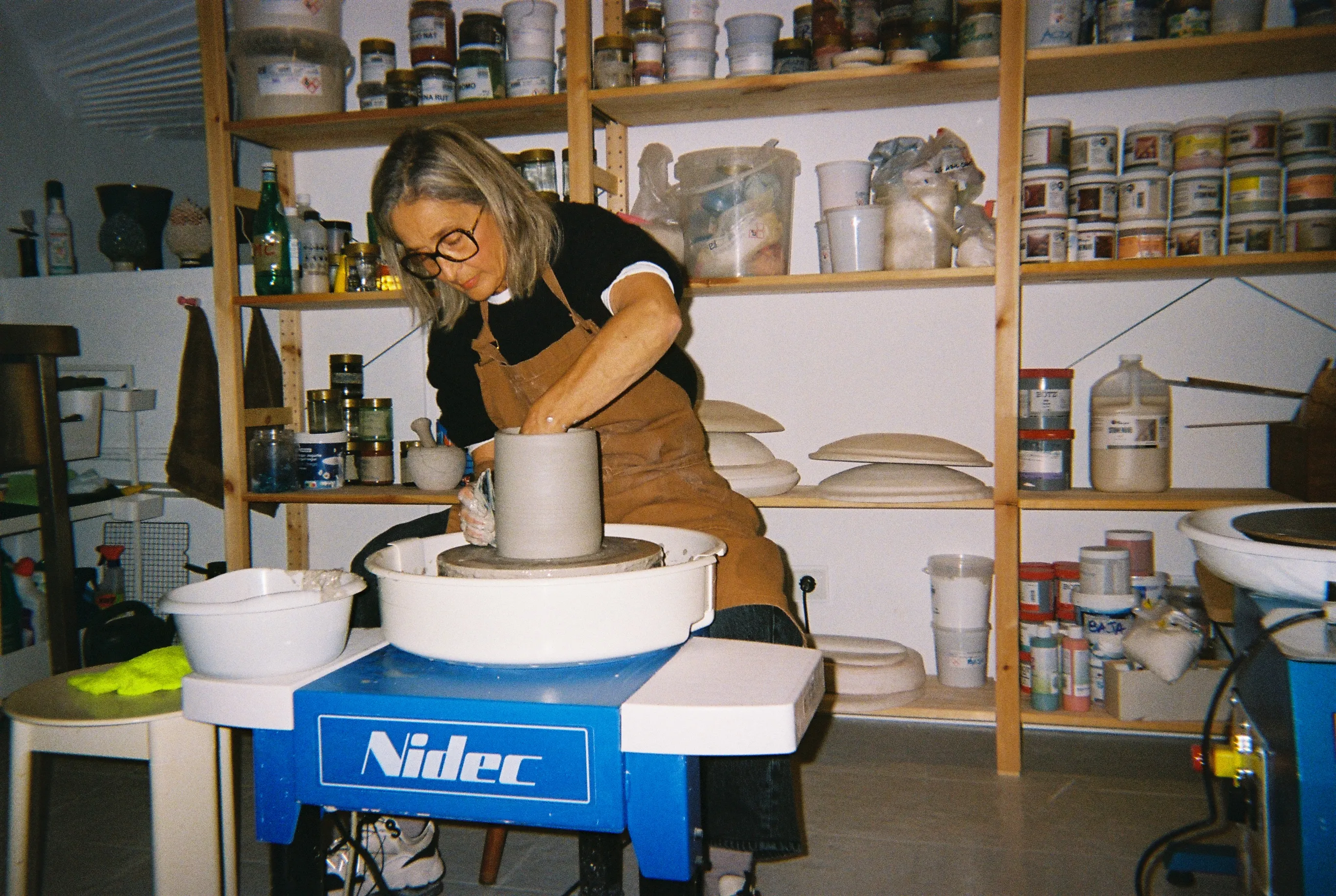 Laura levanta un cilindro de barro en el torno, rodeada por las estanterías de esmaltes del estudio